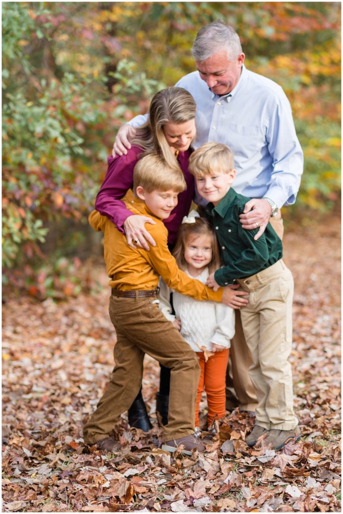 Family of 5 in jewel tone outfits hug with beautiful fall leaves in the background during fall family photos at Sims Lake Park. 