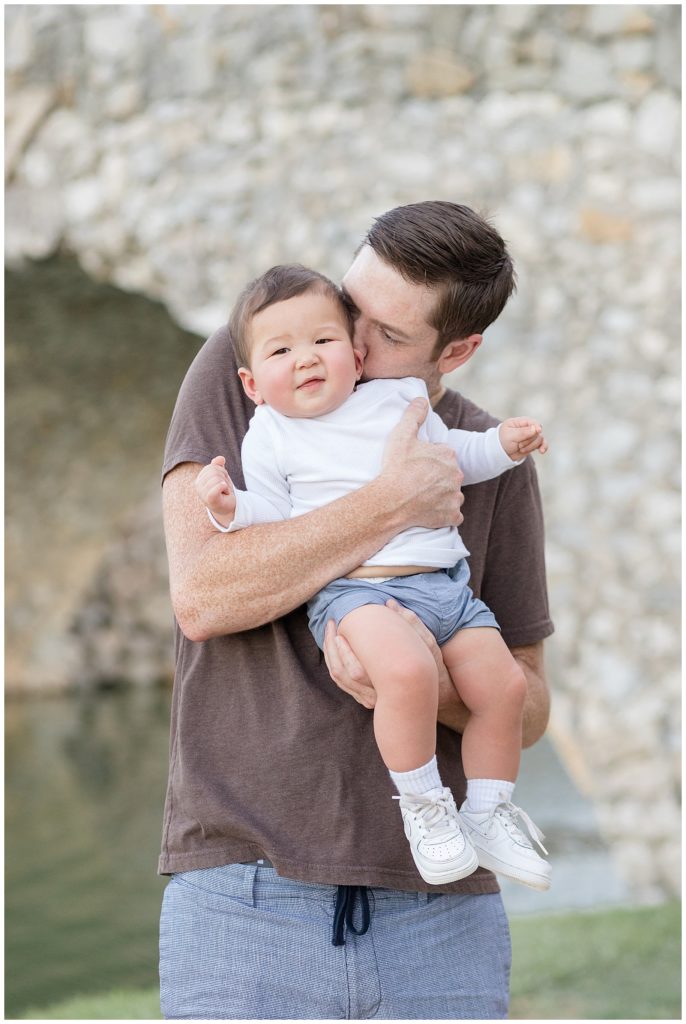 Dad kisses 8 month old son to get him to smile for camera of Wisp + Willow Photography Co. camera.  Father and son coordinate in white, denim, and brown for family photography session in McKinney, TX.