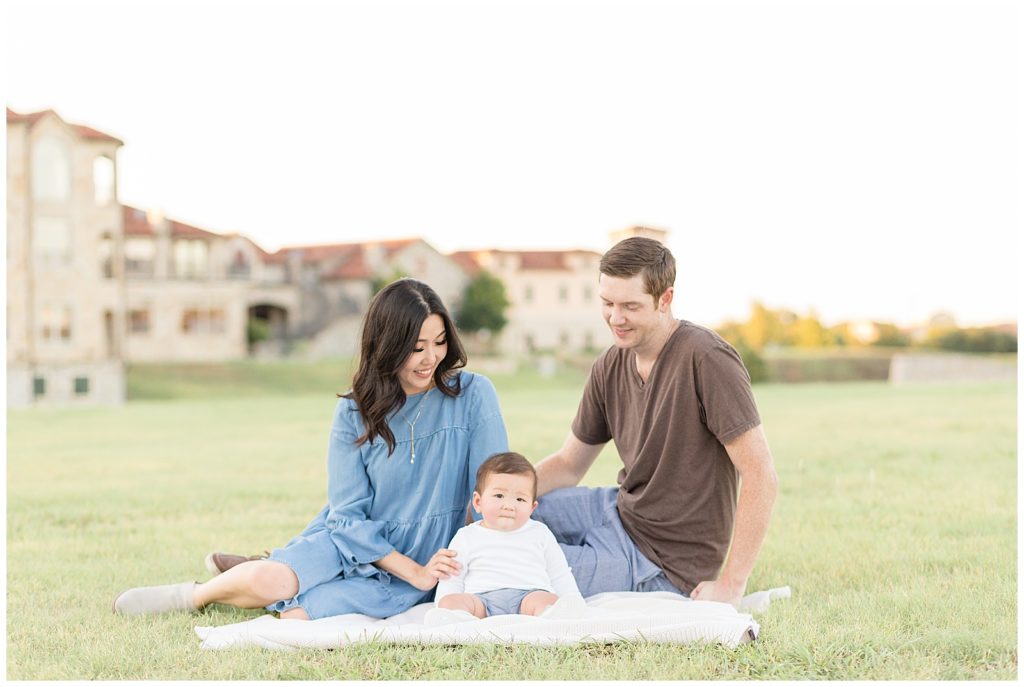 Family of 3 sit in field at Adriatica Village in McKinney, TX with mom and dad looking down at baby boy for their family photography session.