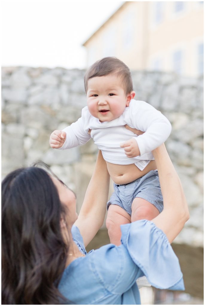 Mom holds baby boy up in the air as we smiles at the camera of Wisp + Willow Photography Co. camera during their fall family session in McKinney, TX at Adriatica Village.