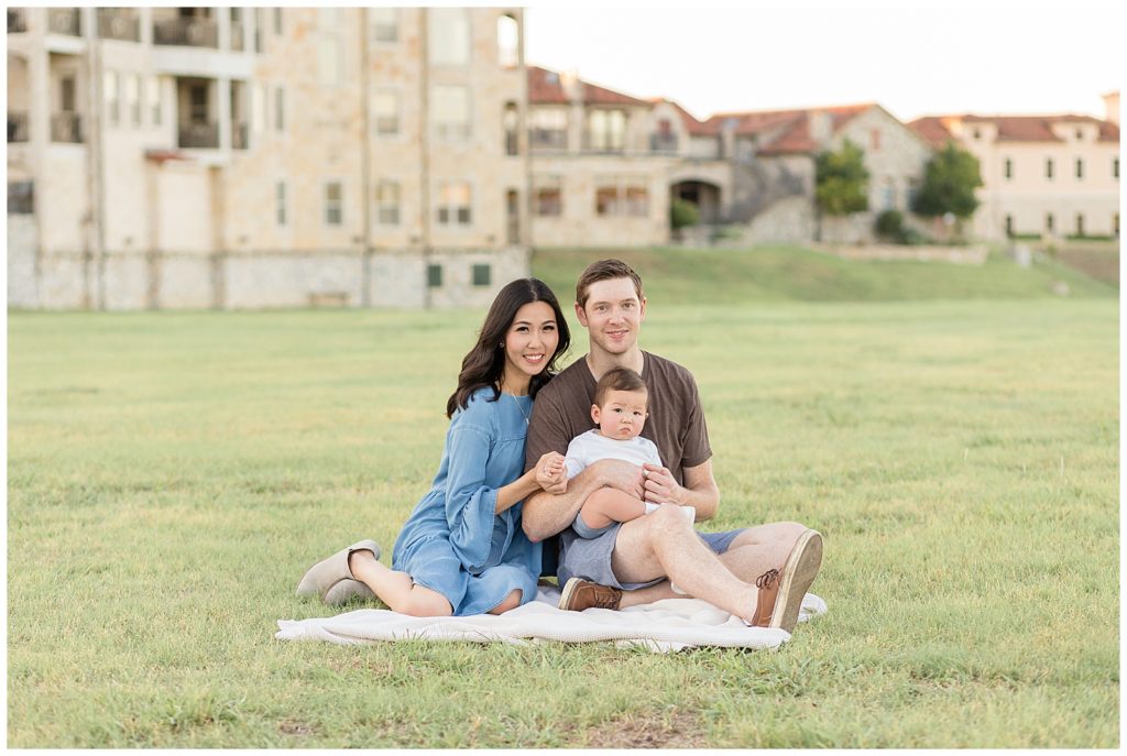 Family of 3 smile at Wisp + Willow Photography Co. for their fall family session at Adriatica Village in McKinney, TX.  Mom wears a denim dress and baby and dad coordinate outfits wearing a mix of denium, white, and brown clothes.