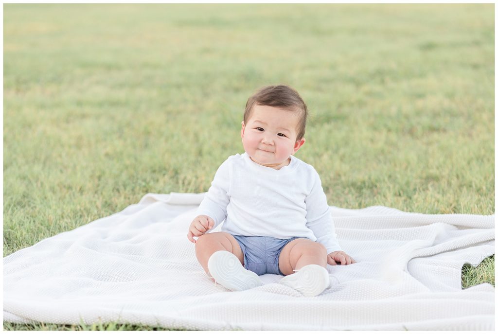 Sweet 8 month old baby boy smiles at Wisp + Willow Photography Co. camera as he sits in field at Adriatica Village in McKinney, TX.  Baby boy wears denim shorts, white shirt, and white shoes!