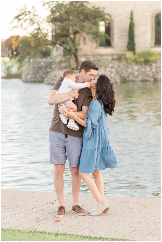 Mom and Dad share kiss as baby boy reaches for mom at Adriatica Village in front of the water.  Photography taken by McKinney, TX family photographer, Wisp + Willow Photography Co.