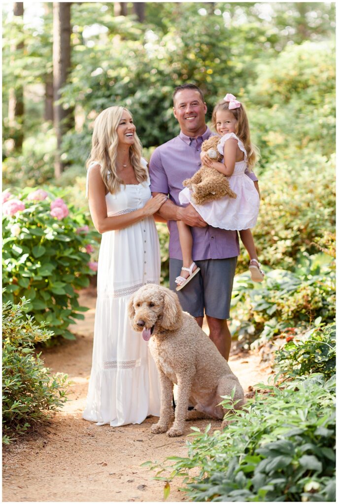 Family of 3 and their goldendoodle stand in the Azalea Gardens in Raleigh, NC wearing coordinating outifts of pink, white, and purples colors during their family photography session with Wisp + Willow Photography Co.