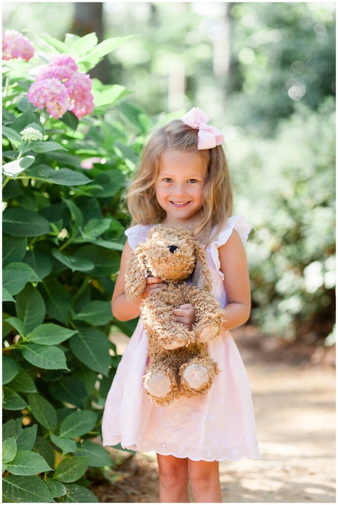 Little girl stands in Azalea Gardens in Raleigh, NC with her stuffed dog animal with pink hydrangeas in the background.