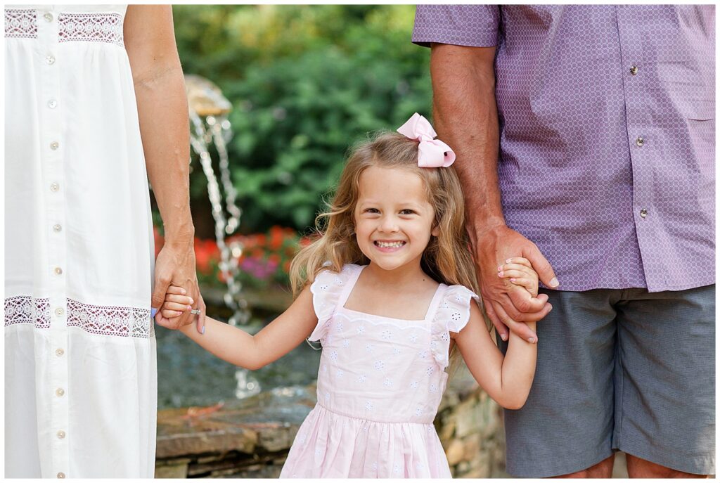 Sweet little girl wears pink eyelet dress and pink bow as she stands holding her parents hands during her Azalea Gardens family photography session in Raleigh, NC.