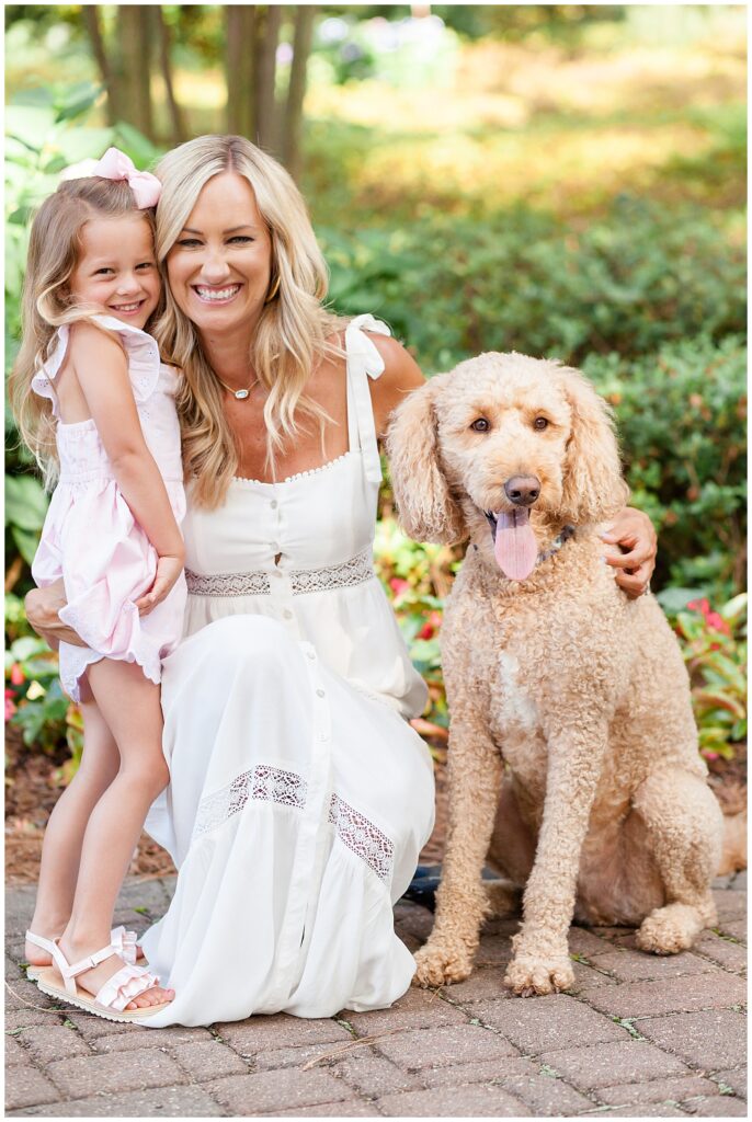 Mom and daughter wear white dress and pink eyelet dress for the little girl as they smile with their goldendoodle puppy during their Azalea Gardens Family Photography session in Raleigh, NC.