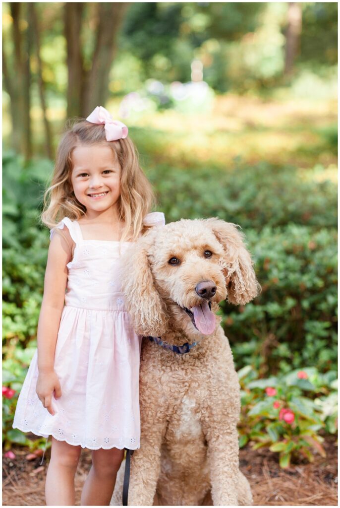 Little girl wears pink eyelet dress with pink bow while smiling during her family photography session in Raleigh, NC with her goldendoodle pup.