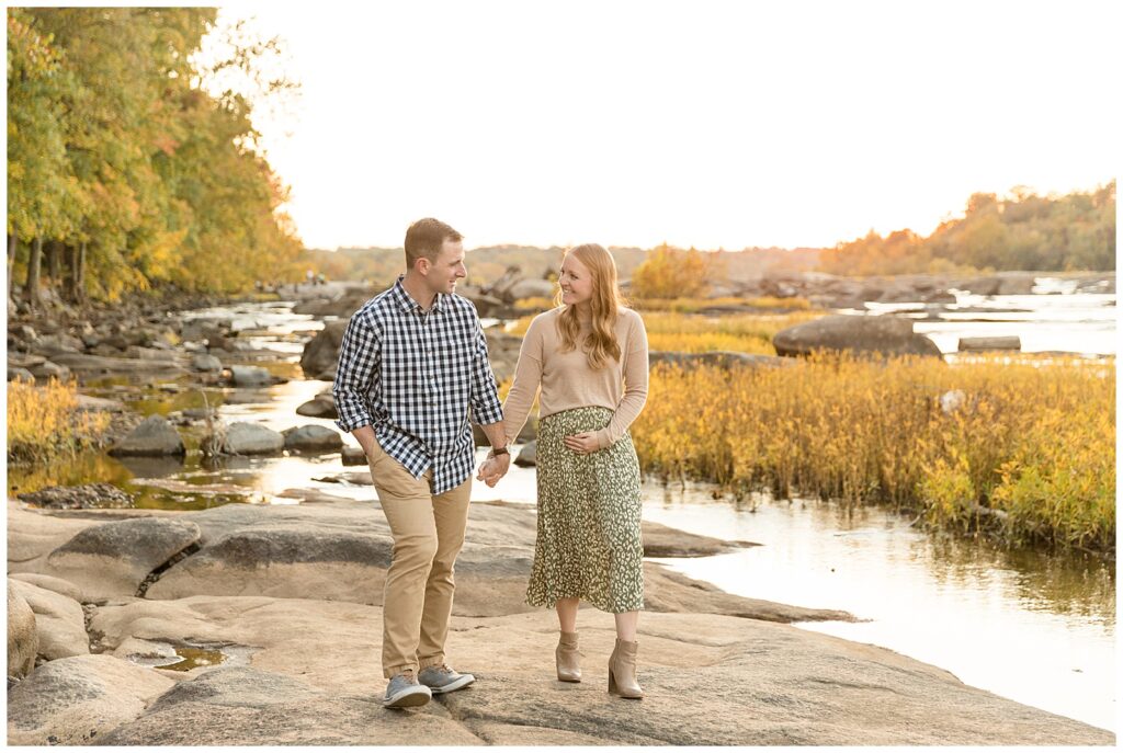 Couple walks along rocks of Belle Isle in front of Richmond family photographer, Wisp + Willow Photography Co. camera as mom to be holds belly to share exciting baby announcement.
