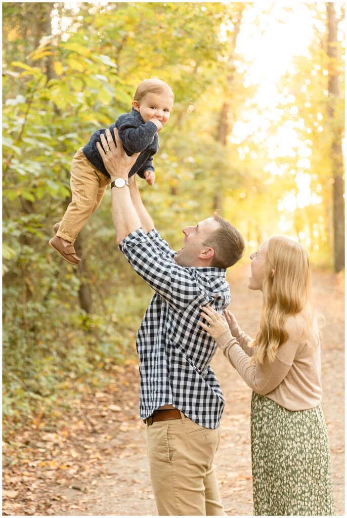 Dad holds toddler son up in the air as boy smiles at camera and mom and dad smile joyfully up at their son during their family session with Wisp + Willow Photography Co.