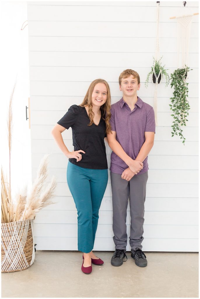 Sister and brother stand in front of white farmhouse wall with green plants and beach seagrass in McKinney, TX photographed by Wisp + Willow Photography Co.