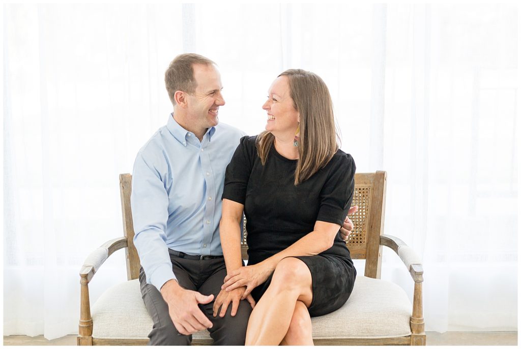 Married couple and mom and dad lovingly look at each other and laugh in studio with sheer white curtain behind them and they sit in wicker farmhouse chair photographed by Wisp + Willow Photography Co. in McKinney, TX.