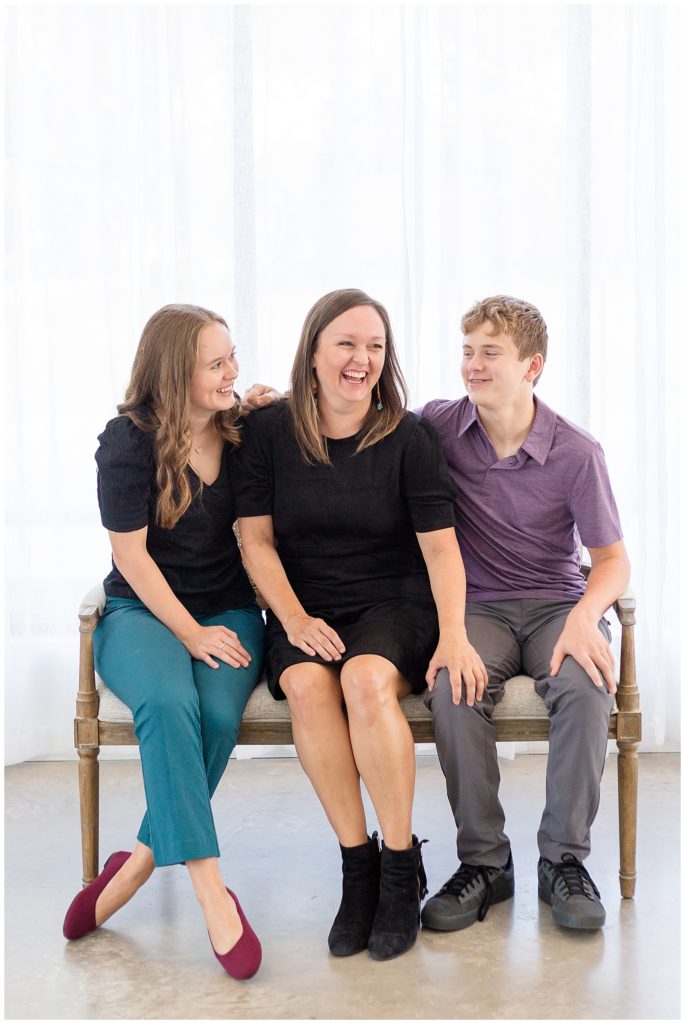 Mom sits with 2 kids a daughter and a son as she laughs and kids look at her smiling.  A joyful image taken in McKinney, TX at Lemon Drop Studios photographed by family photographers Wisp + Willow Photography Co.