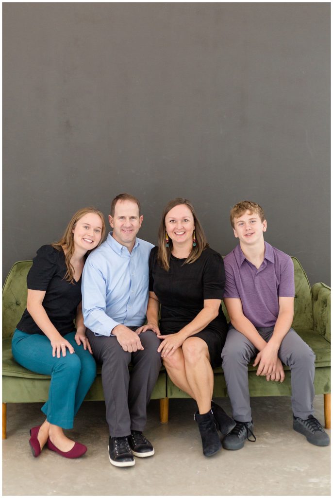 Dad, Mom, daughter, and son smile at camera in studio at Lemon Drop in McKinney, TX.  They are sitting on a green, velvet couch with a dark gray wall behind them.