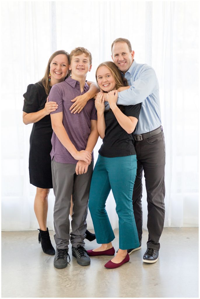 Family bear hugs in family portrait session in McKinney, TX at Lemon Drop Studios.  They coordinate with black, purple, blue, grey, and teal colors in front of studio window with white curtains.