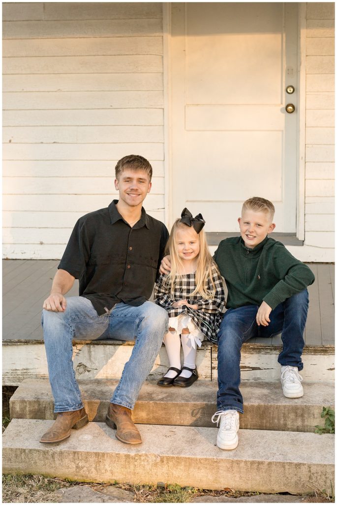 Siblings sit on old porch house steps as they smile for the camera.  Two older brothers and younger sister smile for their updated family Christmas card.