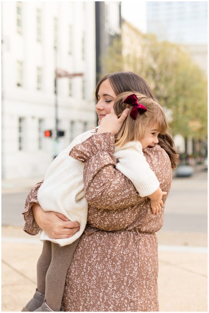 Daughter hugs mom during their mini session in Raleigh, NC wearing a burgundy bow and coordinating brown tights and floral brown dress.  See more of Wisp + Willow Photography Co. work on the blog now! 