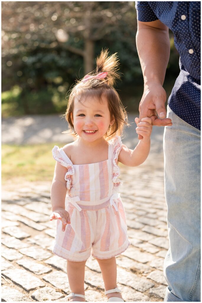 Little girl holds Dad's finger as she smiles at the camera of Wisp + Willow Photography Co. and wears linen striped romper that has purple, coral, and white and cream stripes.