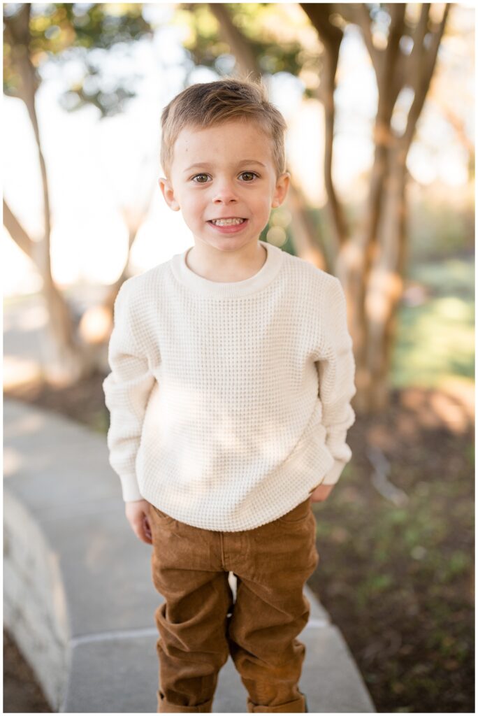 Little boy cheeses at camera of Richmond, Virginia family photographer, Wisp + Willow Photography Co. wearing a cream, waffle knit shirt and brown jeans.