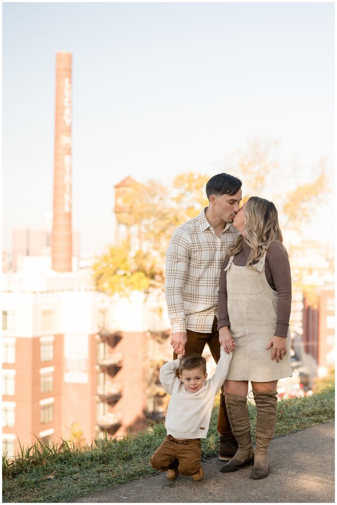 Mom and Dad share a kiss in front of landmark at Libby Hill Park as they hold hands of son in front of them.  You can see more work from Wisp + Willow Photography Co. on the blog now!