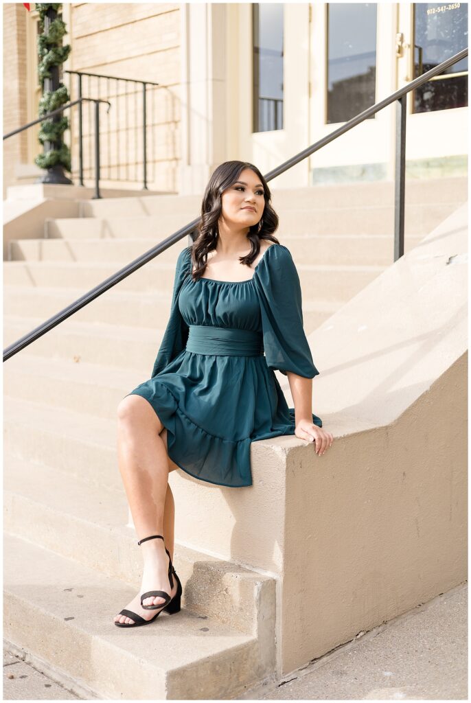 Senior session in Downtown, McKinney has girl wearing a gorgeous, dark green dress with puff sleeves as she sits on steps outside of store.