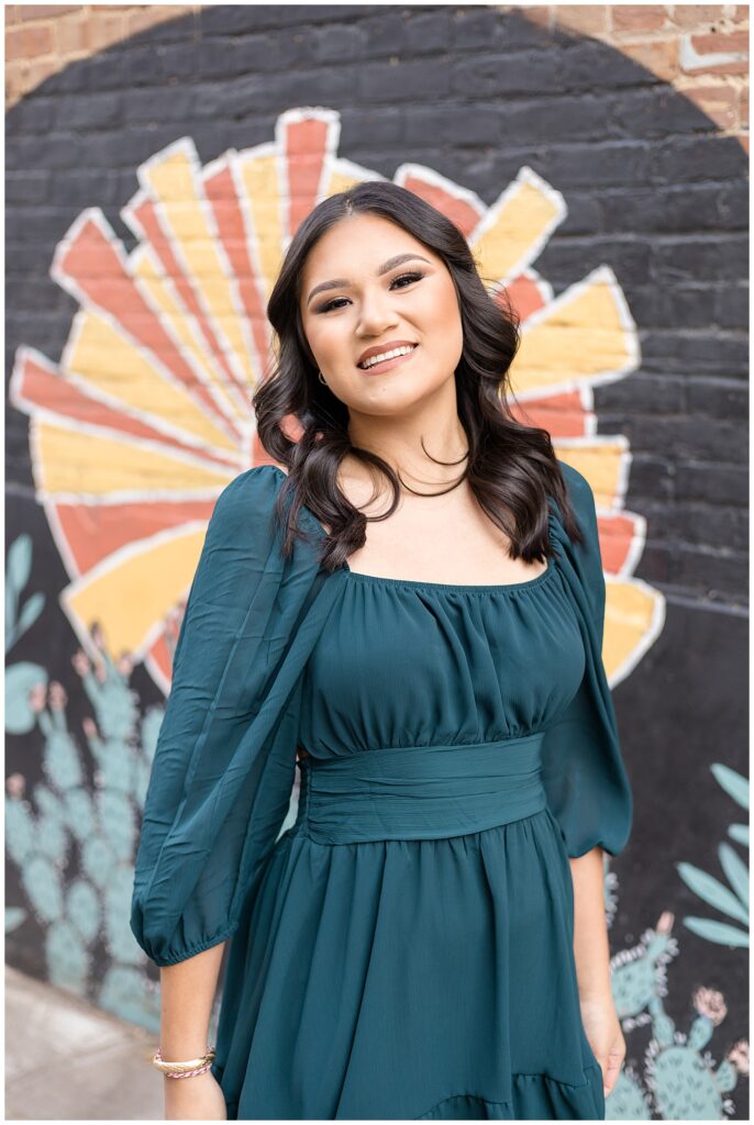 Girl wears dark green dress in Downtown, McKinney, TX, square standing in front of mural with a sunshine for her senior portrait session.
