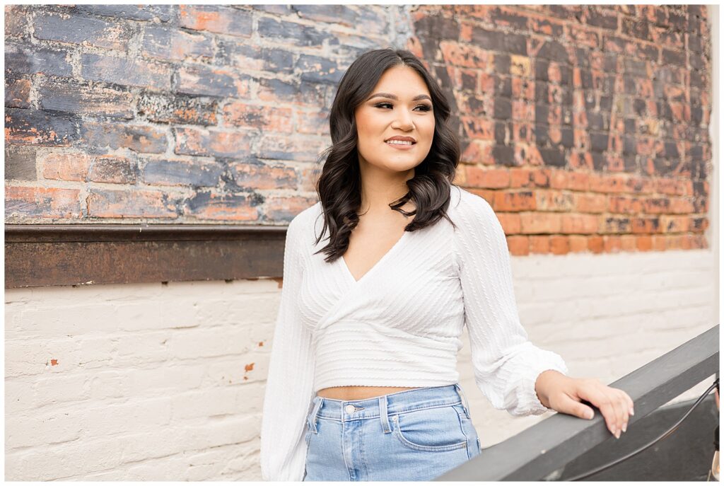 Senior session in Downtown Square in McKinney, TX captures girl standing in front of brick building wearing a white crop top and jeans as she looks off into the distance.