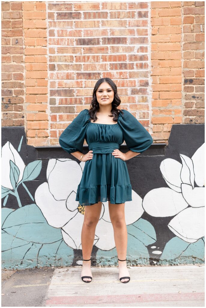 Senior session in downtown McKinney, TX has senior girl standing in front of a brick building with a floral mural painted in black, teal, and white.