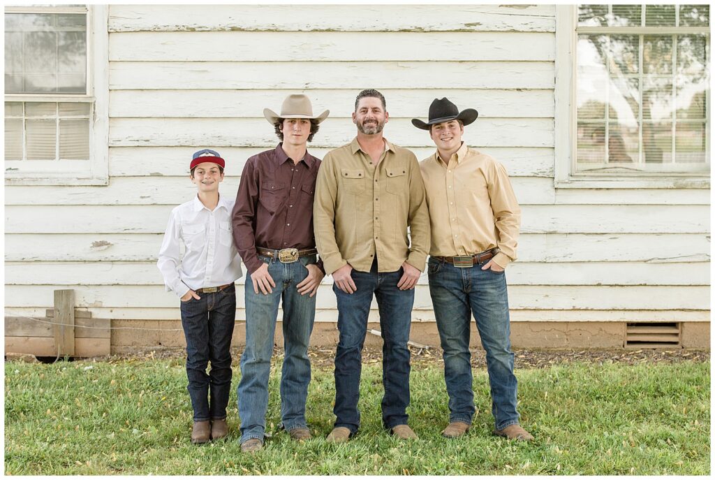 Dad and his three cowboy sons stand in front of barn with their thumbs in their pockets wearing a mix of tan, white, and burgundy shirts.