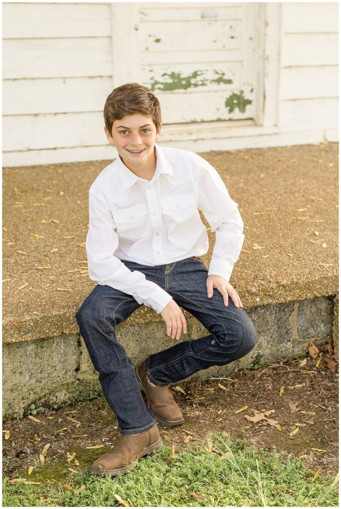 Youngest pre-teen son sits on steps of barn and smiles at Franklin family photographer, Wisp + Willow Photography Co. wearing a white shirt, jeans, and cowboy boots.