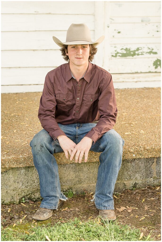 Teenage boy sits on the steps of barn in Franklin, TN wearing a cowboy hat, brown shirt, and jeans during their family session with Wisp + Willow Photography Co.
