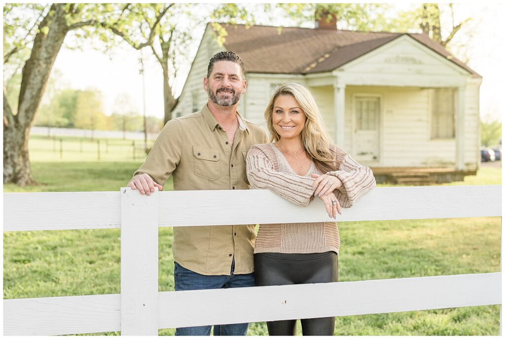Husband and wife stand behind white picket fence at Halinsdale Farm in Franklin, TN for their family mini-session with photographers, Wisp + Willow Photography Co.