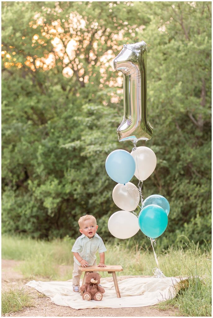 Milestone photo session in Plano, TX captures first birthday of boy who stands with his hands on a wooden stool with his teddy bear in front and blue and white balloons and one silver with the number one.