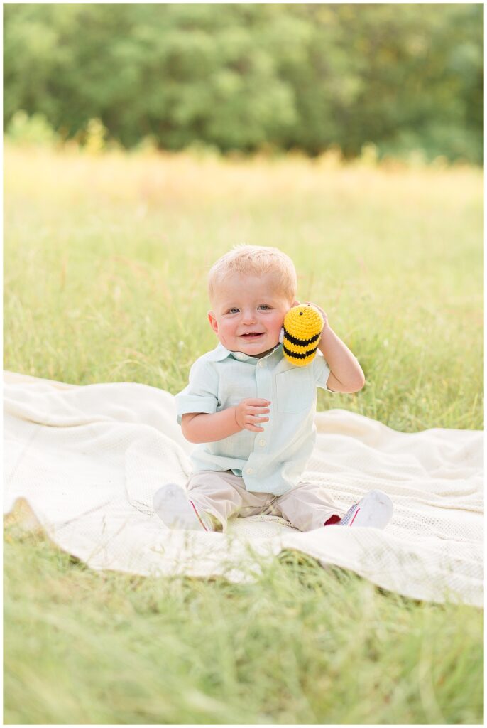 Little boy sits in field on blanket wearing a green shirt and khaki pants while he holds a yellow and black bumble bee toy to his cheek and ear.