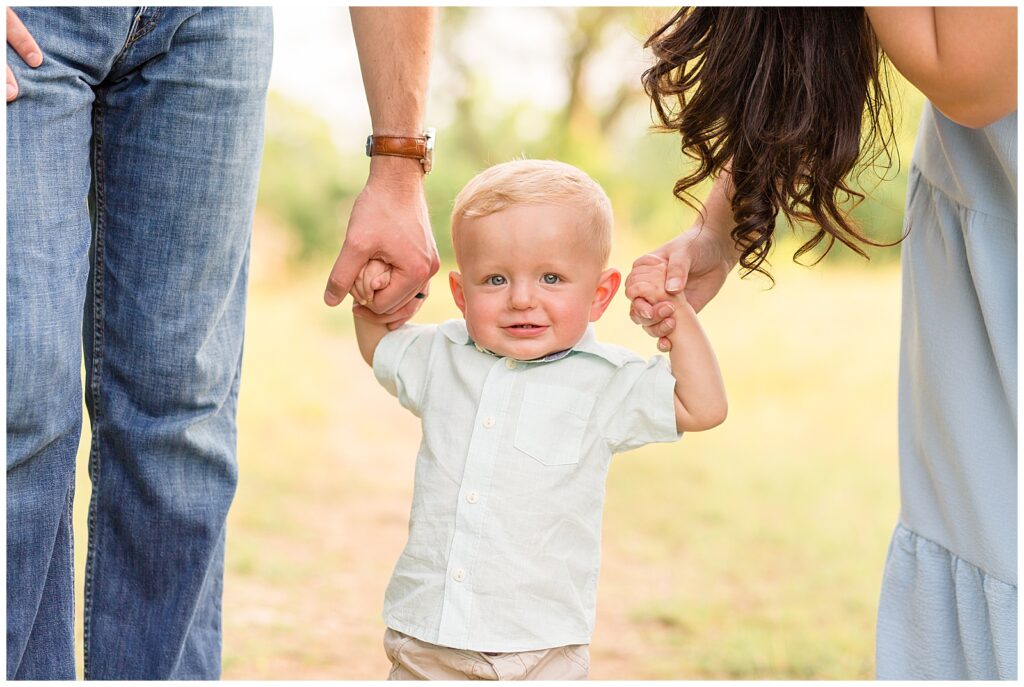 1 year hold boy holds hands of mom and dad as he smiles at camera with blonde hair, blue eyes, green button down shirt and khaki pants.