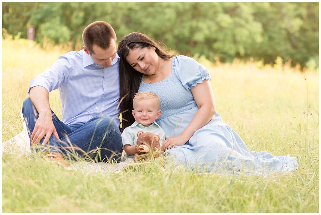 Family of 3 sit in field with 1 year old boy as he holds his brown teddy bear and smiles at camera.