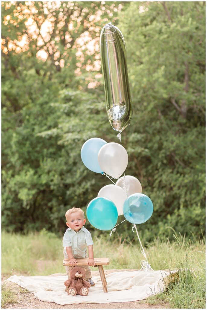 1 year old milestone photo shoot captures little boy who stands behind a wooden stool where his brown bear is sitting in front of and blue and white balloons are beside them.