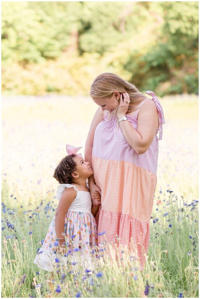 Mother and daughter stand in field of flowers looking at each other smiling sharing a tender moment.