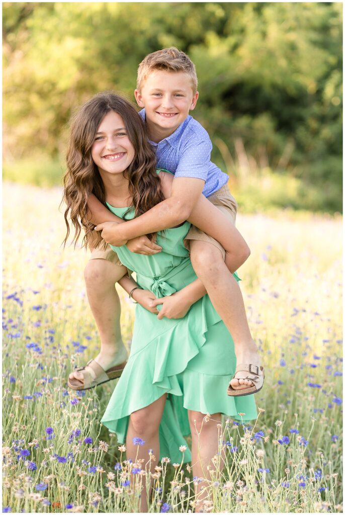 Sister carries little brother on back as they smile for their camera during their family mini photography session.