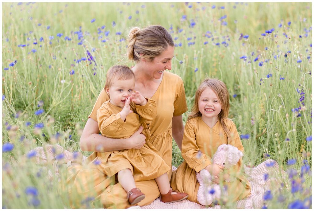 Mom sits with two daughters, little girl and baby, in a wildflower field wearing matching mustard yellow dresses. Texas family photographer, Wisp + Willow Photography Co.  capture cherished moment.