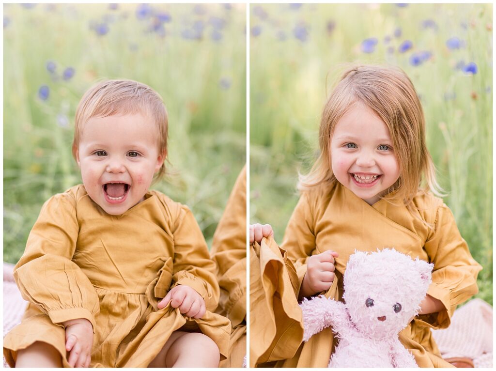 Little girl smiles happily in wildflower field holding her pink stuff animal for their family photography mini session.  Baby sister also smiles big for the camera in her mustard yellow dress.