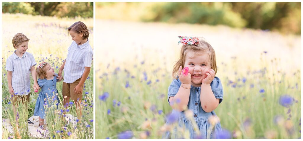 Little girl holds holds her cheeks as she holds a bright pink flower and wears a denim dress and floral bow in a field of flowers.  Two brothers and little sister stand holding hands looking at each other and smile.