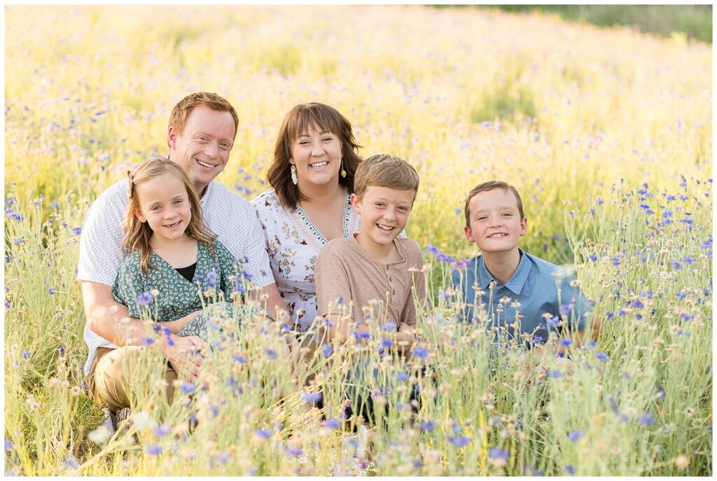 Family of 5 sit in wildflower field wearing coordinating outfits of blue, green, white, and light brown and smile for the camera of Wisp + Willow Photography Co.