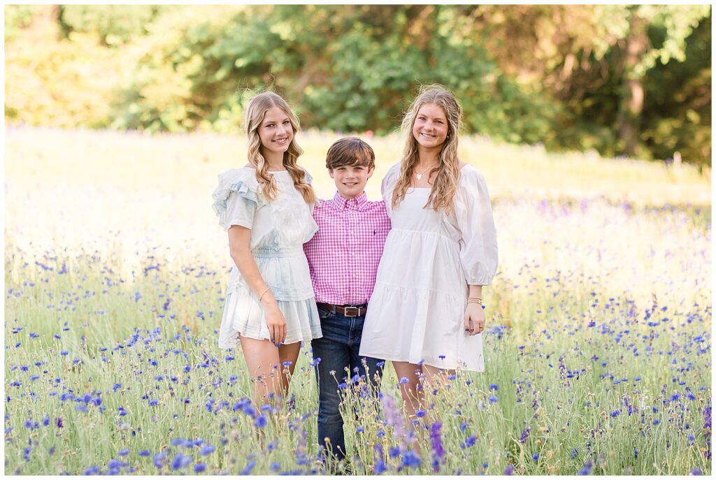 Two older sisters wearing a white dress and the other a light white and blue tie-dye romper, stand with little brother who wears a pink plaid shirt and jeans in a wildflower field in TX.