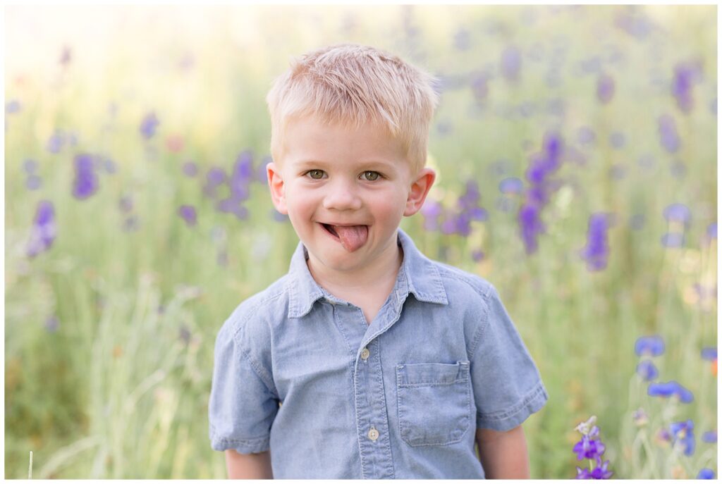 Cute little blonde boy wears denim shirt and sticks his tongue out at camera during his wildflower family mini photography session.  Texas family photographer, Wisp + Willow Photography Co. couldn't have captured a cuter shot!