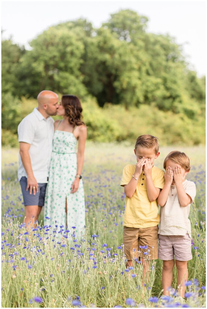 Mom and dad kiss while two sons are the in the forefront of the shot covering their faces with their hands to not see their parents kiss during a texas family photographer session with Wisp + Willow Photography Co.