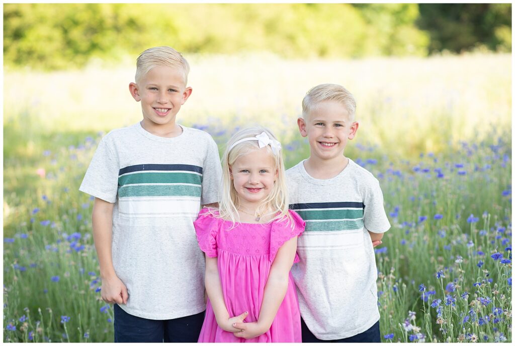 3 little blonde siblings, two older brothers and one younger sister smile at camera of Wisp + Willow Photography Co., Texas family photographer, during their wildflower mini session.