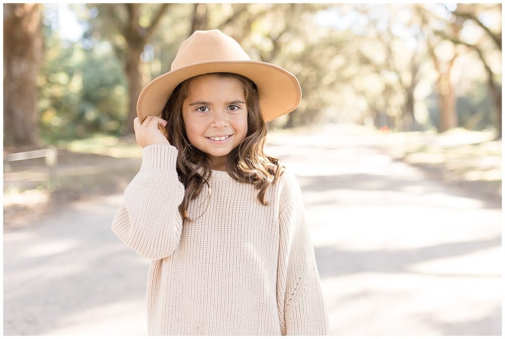Little girl wears influencer hat and a cream sweater while standing on pathway surrounded by Spanish moss during fall mini session.