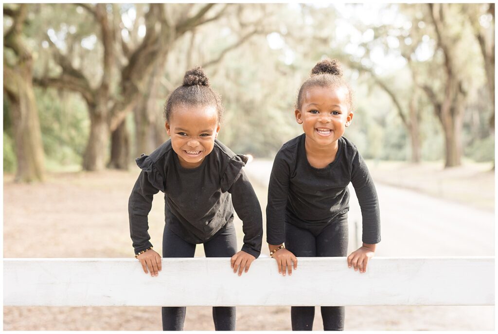 Twin toddler sisters smile happily at camera of Savannah family photographer, Wisp + Willow Photography Co. as they stand on picket fence with Spanish Moss surrounding them.