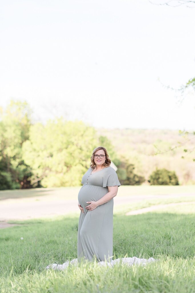 McKinney Maternity Portrait Session at Erwin Park has mom-to-be wearing a muted mint colored dress while she holds her belly and smiles at the camera of Wisp + Willow Photography Co.